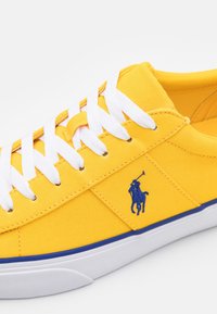 Gul canvas sneakers med vita snörningar, en vit gummisula med blå kant och en blå broderad polo-spelarlogo på sidan.