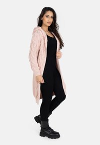 Long cardigan en tricot rose avec une capuche, présentant un motif en maille torsadée, porté sur un ensemble noir ajusté, associé à des bottes noires à lacets.
