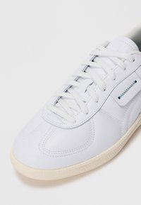 Hvite lær-sneakers med kremfarget såle, hvite lisser, søm detaljer og en liten klaff med blå sikksakk-søm på siden.