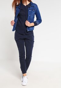 Denimjakke i medium blå farve parret med en marineblå bluse og mørk marineblå bukser, suppleret af hvide sneakers. Glatte teksturer og klassisk design.