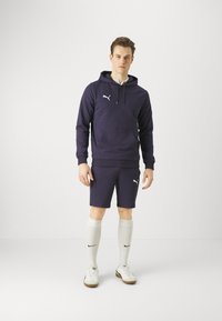 Marinefarbener Kapuzenpullover mit Kängurutasche, kombiniert mit marinefarbenen Shorts. Weiße Logos auf beiden. Das Model trägt hellgraue kniehohe Socken und weiße Sneakers.