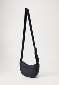 Svart tøy crossbody-veske med en buet form, justerbar stropp og en frontlomme med glidelås. Har en jevn tekstur og et minimalistisk design.
