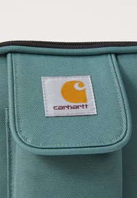 Geantă din material teal cu un finisaj texturat, având un eticheta alb�ă cu un logo portocaliu și text roșu. Închidere cu fermoar în partea de sus.