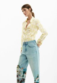 Cămașă galbenă în carouri, cu nasturi și un design cu volane, asortată cu blugi deschis la culoare, albastru, având pe piciorul stâng o scenă costieră colorată imprimată.