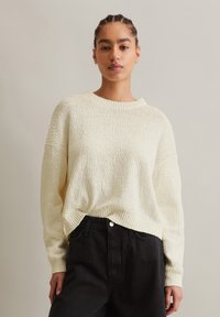 Cremfarbener Pullover mit strukturierter Textur, weitem Ausschnitt, überschnittenen Schultern und gerippten Bündchen, kombiniert mit hochtaillierten schwarzen Jeans.