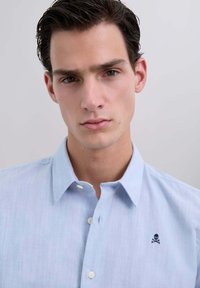 Camisa de botones azul claro hecha de una tela texturizada, con un pequeño logo bordado de calavera y tibias cruzadas en el pecho.