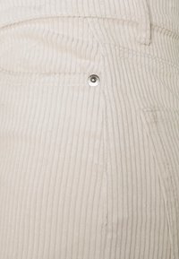 Tissu en velours côtelé beige clair avec des rainures verticales, doté d'un bouton-pression argenté et d'un détail de poche latérale. Surface texturée.