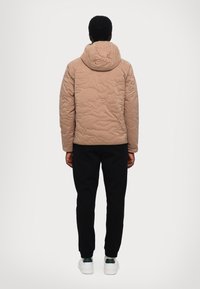Quilted beige jakke med hætte, der har en ujævn tekstur. Matcher med sorte bukser og hvide sko, set fra ryggen.