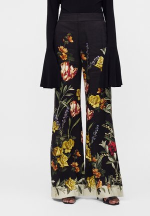 Pantalon noir à jambes larges avec un motif floral coloré comprenant des roses jaunes, des tulipes et des feuilles vertes, associé à des sandales à talons noires.
