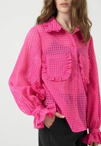 Vrouw in een doorzichtig fuchsia blouse met een geblokt patroon, volants aan de kraag, kantjes aan de zakken en opbollende, gerimpelde manchetten, gecombineerd met zwarte broek.