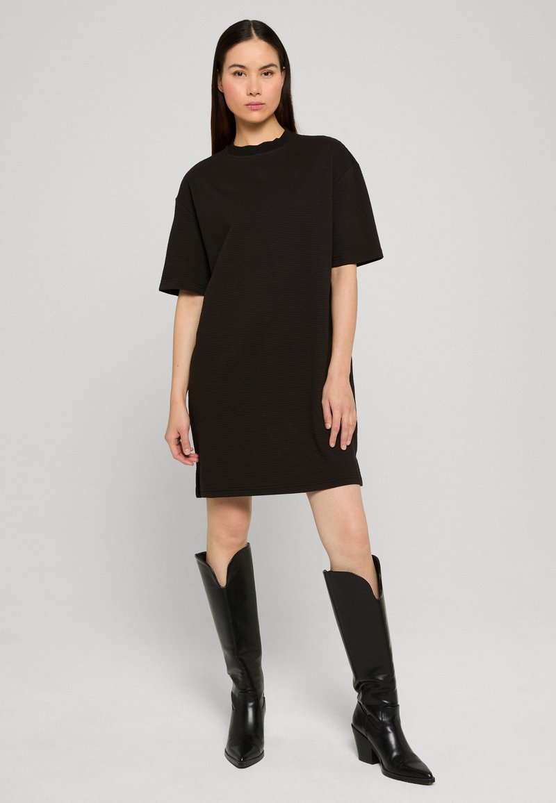 Femme portant une robe noire ample à manches courtes, associée à des bottes noires à talons montant jusqu'aux genoux, debout devant un fond clair uni.