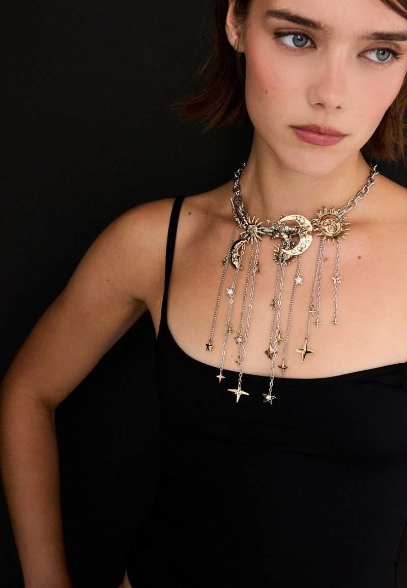 Jeune femme aux cheveux courts bruns portant un haut noir sans manches et un collier doré à thème céleste avec des breloques soleil, lune et étoile avec des chaînes.