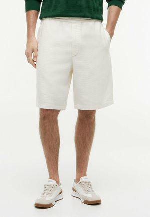 Mann trägt weiße, strukturierte Shorts mit Taschen, weiße Sneakers mit braunen Sohlen und ein grünes Langarmhemd.
