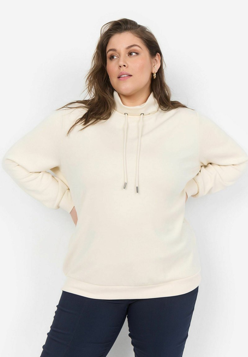 Sudadera de color crema con un cuello alto y cordones, con una textura suave y un corte relajado. Llevada con pantalones oscuros.