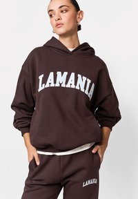 Hanorac maro cu glugă, cu inscripția "LAMANIA" imprimată alb pe piept. Croială lejeră, cu manșete rib și textură moale.