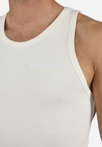 Hvit, ribbestrikket tanktop med rund hals, med en brodert merkevarelogo i lys blå på forsiden. Mykt, strukturert stoff.