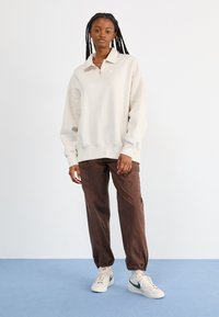 Ανοιχτό μπέζ φούτερ με μισό φερμουάρ στο γιακά, συνδυασμένο με καφέ cargo παντελόνι και άσπρα sneakers με πράσινο σήμα. Ελάχιστη ορατή σήμανση.