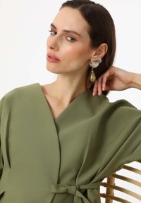 Olive green wrap top made of smooth fabric, featuring a deep v-neck, wide sleeves, and a tie detail at the waist. Gold and white earrings.
