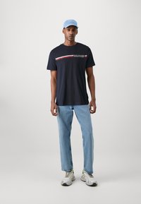 Tricou navy cu mâneci scurte, decorat cu dungi orizontale albe și roșii și broderie "HILFIGER" pe piept; asortat cu blugi ușor albaștri.
