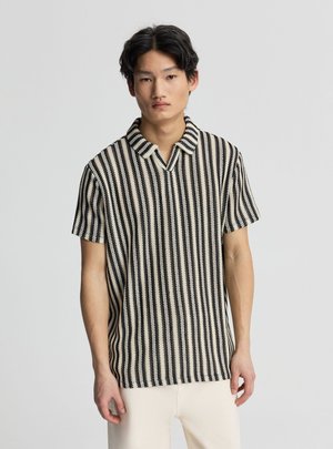 Giovane uomo con capelli corti e neri che indossa una maglia a maniche corte a righe verticali bianco e nero e pantaloni chiari, in piedi davanti a uno sfondo grigio.