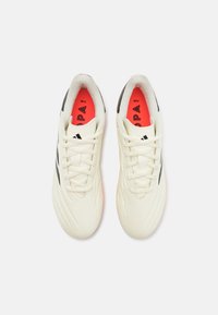 Botas de fútbol de material sintético blanco con rayas negras. Presentan una superficie texturizada y plantillas en rojo brillante con detalles del logo.