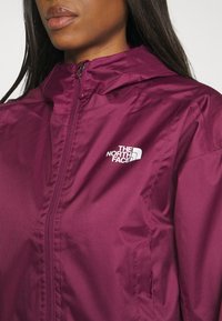 Bordowa, lekka kurtka z zamkiem błyskawicznym z przodu, kapturem i białym logo The North Face na lewej piersi. Gładka, błyszcząca faktura.