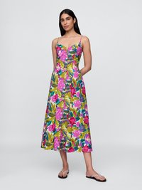 Eine Frau trägt ein ärmelloses, knielanges Hemdkleid mit bunten rosa und grünen Blumenmustern und posiert vor einem schlichten hellen Hintergrund.
