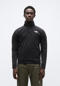 Μαύρο ενδιάμεσο στρώμα από fleece με γιακά με φερμουάρ στο ένα τέταρτο, λεπτομέρειες υφής στο ύφασμα και λογότυπο στο στήθος, συνδυασμένο με ελαφριά πράσινα παντελόνια.