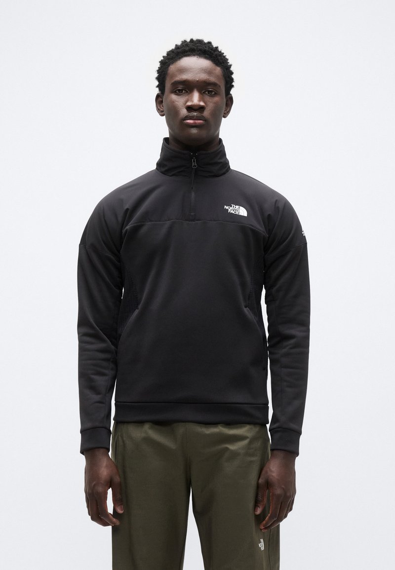 Μαύρο ενδιάμεσο στρώμα από fleece με γιακά με φερμουάρ στο ένα τέταρτο, λεπτομέρειες υφής στο ύφασμα και λογότυπο στο στήθος, συνδυασμένο με ελαφριά πράσινα παντελόνια.