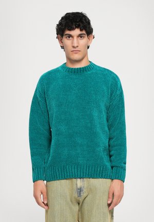 Mann, der nach vorne steht und einen türkisfarbenen gerippten Pullover sowie hellbeige Jeans trägt, vor einem einfachen weißen Hintergrund.