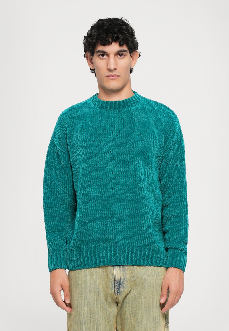 Man standing facing forward wearing a teal ribbed knit sweater and light beige jeans against a plain white background.