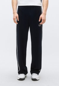 Tmavomodré tepláky s voľným strihom, elastickým pásom, bielymi bočnými pruhmi a malým logom na prednej ľavej strane. Vyrobené z mäkkého materiálu.