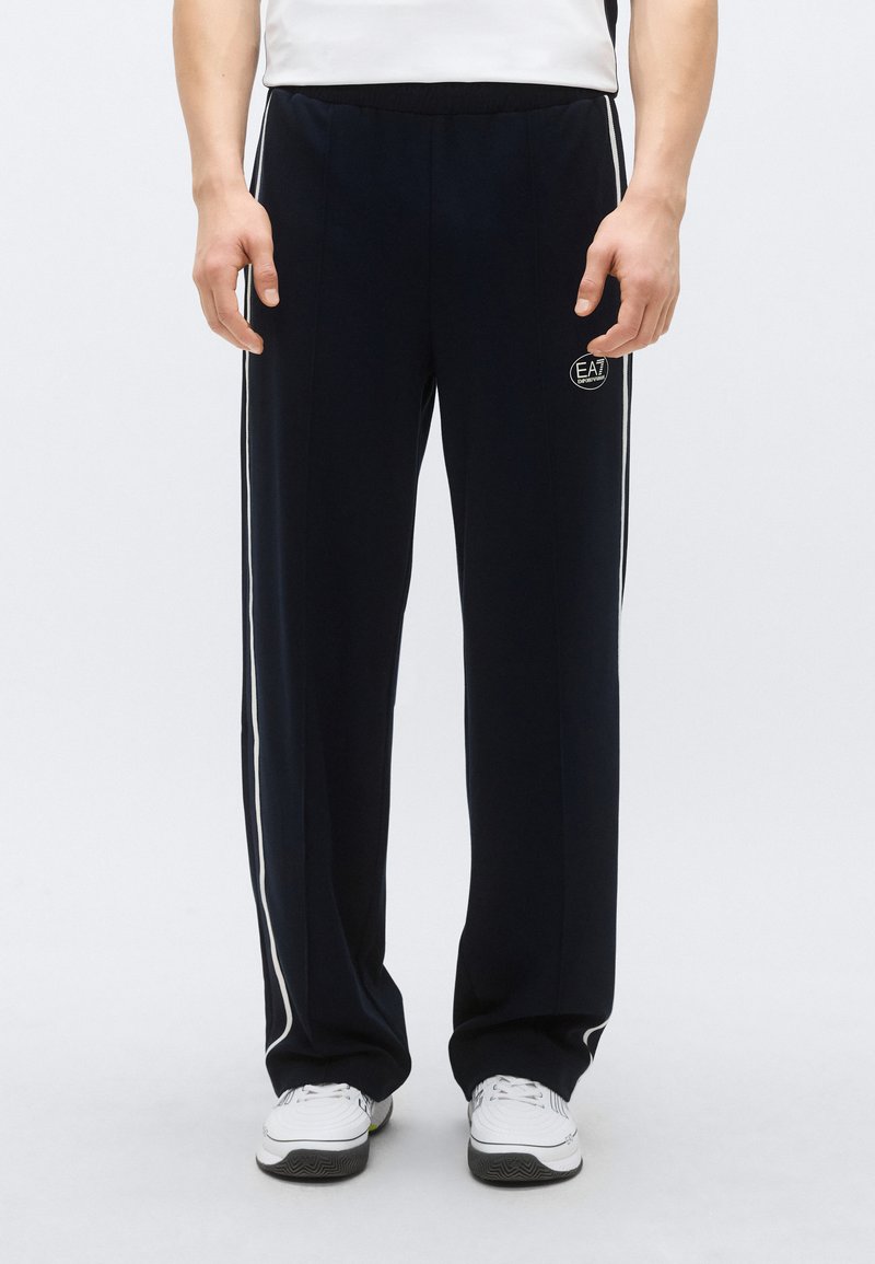 Tmavomodré tepláky s voľným strihom, elastickým pásom, bielymi bočnými pruhmi a malým logom na prednej ľavej strane. Vyrobené z mäkkého materiálu.