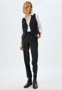 Gilet noir ajusté sur une chemise blanche, associé à un pantalon noir taillé et des bottines noires. Design simple avec des accents de boutons.