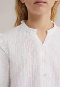 Nahaufnahme einer jungen Person, die eine weiße strukturierte Bluse mit kleinen Knöpfen und einem Rüschensaum trägt.