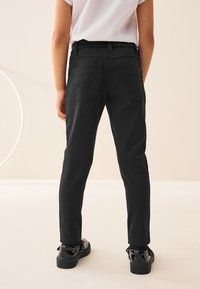 Pantaloni neri slim-fit in tessuto liscio, con due tasche posteriori e passanti per cinture, abbinati a scarpe nere lucide.