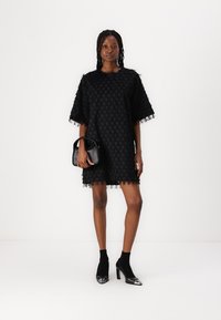 Robe noire texturée à manches courtes, ornée d'un motif et d'une bordure à franges. Associée à des chaussettes noires à la cheville et des chaussures à talons brillantes.