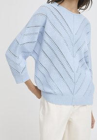 Hellblauer Strickpullover mit lockerer Passform, Zickzackmuster mit offenen Nähten sowie gerippten Bündchen und Saum. Kombiniert mit hellen Hosen.