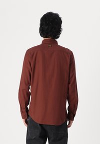 Camisa de manga longa em cor ferrugem com gola com botões. Apresenta um rótulo na parte de trás e punhos com botões. Feita de um tecido texturizado.