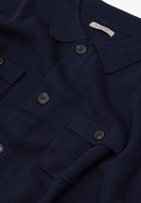Cárdigan de punto azul marino con cuello de solapa, dos bolsillos en el pecho y botones negros. Textura suave con un acabado liso.