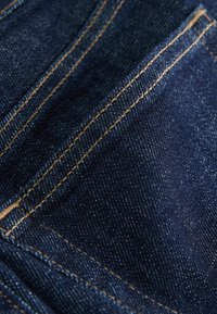 Mørkeblå denimstof med en glat tekstur, som har kontrasterende gyldent syning langs sømmene og lommerne, der viser et klassisk design.