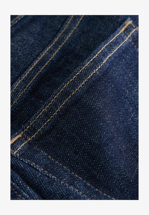 Dunkelblauer Denim-Stoff mit einer glatten Textur, der kontrastierende goldene Nähte entlang der Nähte und Taschen aufweist und ein klassisches Design zeigt.