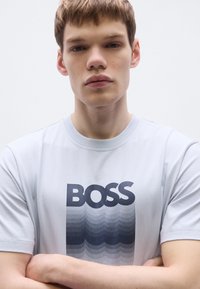 Helehall puuvillane t-särk, millel on tume sinise tooniga julge "BOSS" logo koos gradientse lainemustriga. Lühikesed varrukad ja ümmargune kaelus.