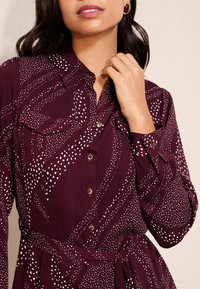 Bordeauxfarbene Hemdblusenkleid mit weißen Punkten, ausgestattet mit Brusttaschen, einem gebundenen Taillengürtel und langen Ärmeln mit geknöpften Manschetten.