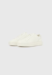 Baskets basses en cuir blanc avec lacets et semelles épaisses en caoutchouc, présentées côte à côte sur un fond gris clair.