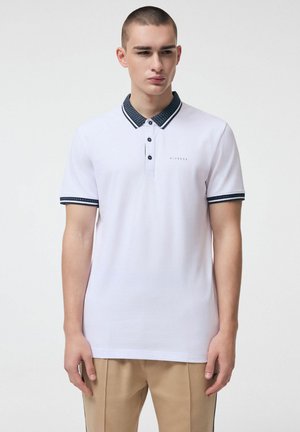 Młody mężczyzna w białej koszulce polo z ciemnym wzorzystym kołnierzykiem i krawędziami rękawów, w beżowych spodniach, stojący na jednolitym tle.