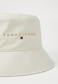 Beige bucket hat made of soft fabric. Features embroidered "TOMMY JEANS" and a small logo with red, white, and blue accents. Circular brim.