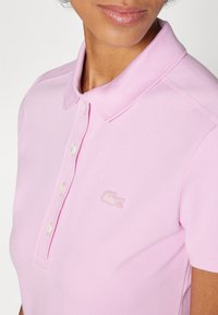 Roze polo majica s teksturiranom površinom, koja ima klasični ovratnik, kopčanje na tri dugmeta i izvezenu oznaku na lijevoj strani grudi.