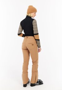 Zwart en tan aansluitend top met mouwtjes in luipaardprint, gecombineerd met tan skibroeken. De broek heeft ritsen bij de enkels en achterzakken. Gebreide muts.