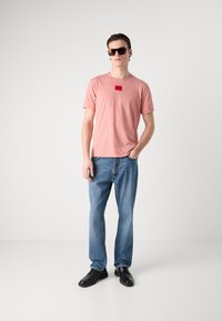 Rózsaszín, rövid ujjú póló piros címkével, kék farmernadrág és fekete cipő. A modellen napszemüveg van, egyik kezével zsebében áll.